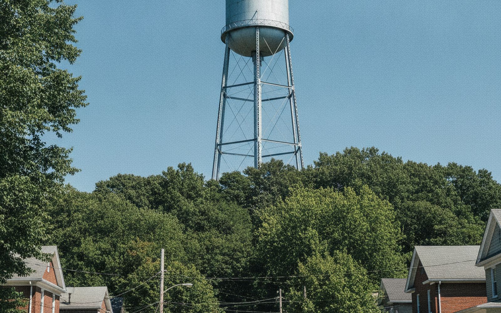 A tall cylindrical water tower rising above summer trees on high ground, seen from a residential street below, blue sky, no signage or lettering visible